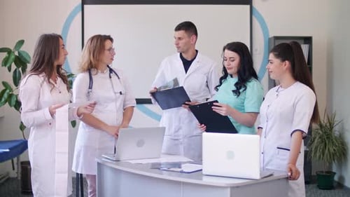 Team of Four Doctors in White Coats Sitting at Table in Hospital and Working With a Laptop