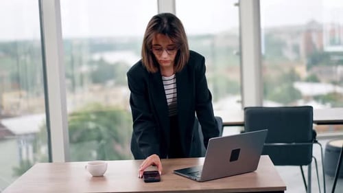 Business lady standing at the desk looks at her smartphone. Woman works on her laptop in the office.