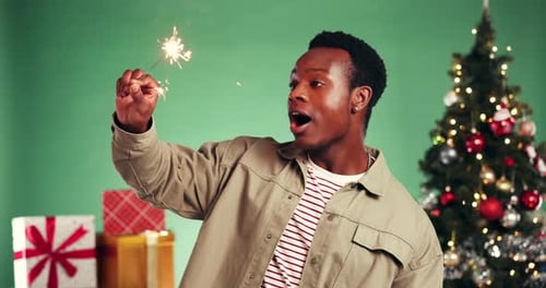 Man, sparkler and celebration with Christmas tree in studio, excited and laugh on green background