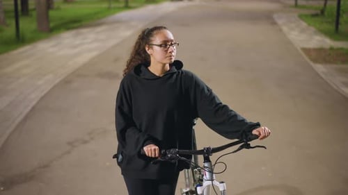 Caucasian Woman Steadying Bicycle On Quiet Park Road At Night Composed Expression Hands On