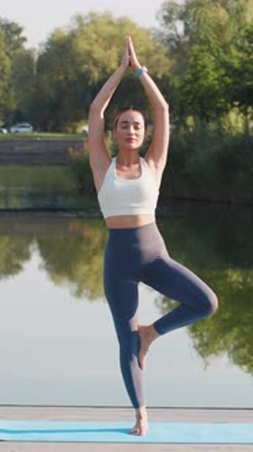 Attraktive junge Sporttrainerin in Sportkleidung, die in Yogabaum-Pose in der Nähe eines Teiches im Park steht