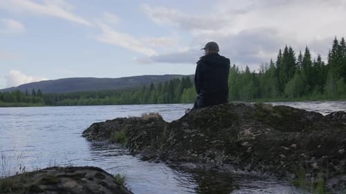 Young man sitting on mossy rock island by wide flowing river, waving to in slow motion