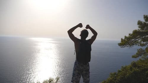 Unrecognizable Hiker with Backpack Standing on Edge of Mountain and Victoriously Raising Hands Male