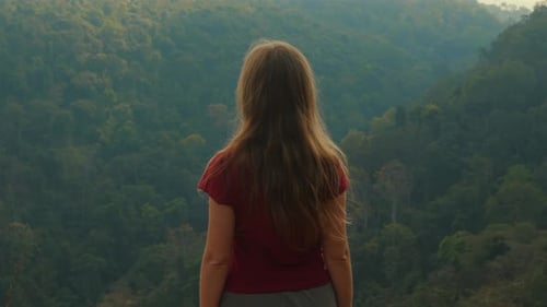 Woman with Long Hair Stands on Cliff Overlooking Lush Green Forest
