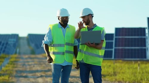 At Sunlight Engineers with Laptop on the Solar Farm in Special Uniform Standing Discuss the
