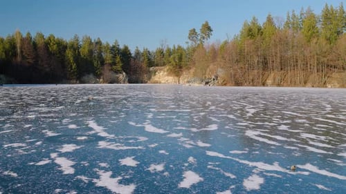 Wide panoramic view of a frozen quarry lake surrounded by pine forest and rocky cliffs. Blue ice