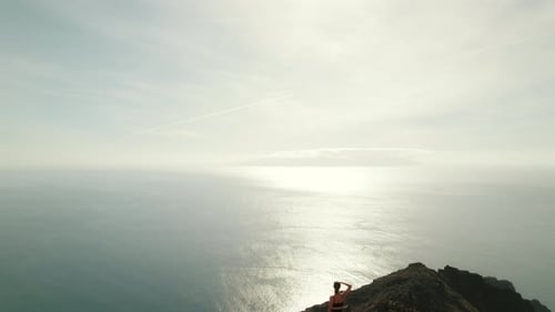 A woman atop a rocky cliff observes the expansive ocean and distant land under a clear sky. Hiker ov