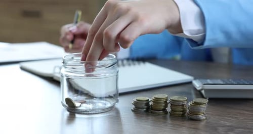Woman putting coins into glass jar while working at wooden table indoors, closeup