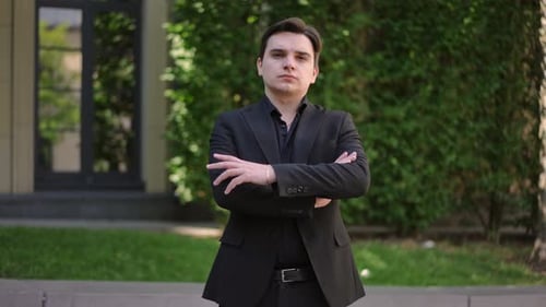 A Serious Young Man in a Dark Suit Poses with His Arms Crossed in Front of Him and Looks at the