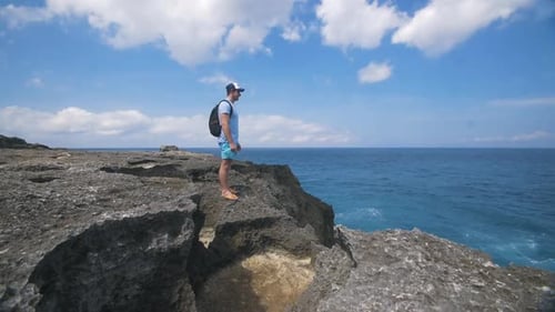 Traveling Man Standing on Edge of Rocks Looking Out at Ocean - Pan Up Reveal.