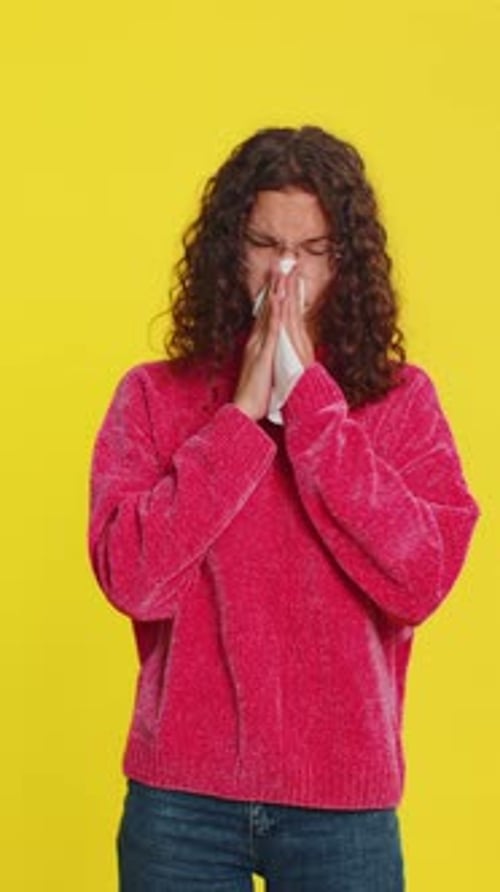 Young Woman Blowing Nose Against Yellow Backdrop