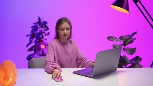 Woman Using Smartphone While Working on Laptop in Colorful Neon Office with Modern Interior Design