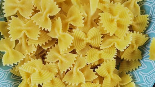 Bow-shaped pasta is poured into a blue ceramic bowl. Top view. Slow motion.