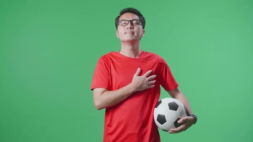 Asian Man With A Ball Putting Hand On His Heart To Respect While Cheering Soccer On Green Screen