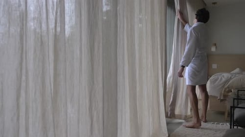 Man in Robe Opens Curtains to Rainy Forest View