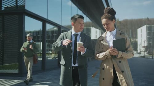 Two Professionals Equipped with Coffee and Folders Discuss Project Progress Amidst Modern Office