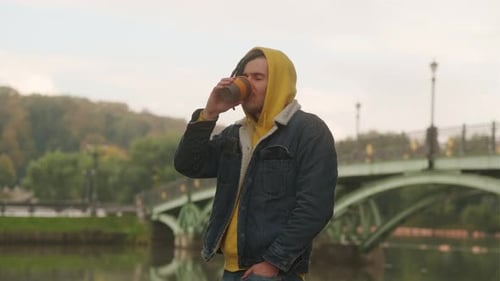 Young Man with Dreadlocks Holding Cup of Coffee Standing Near River Lake on Background of Big Bridge