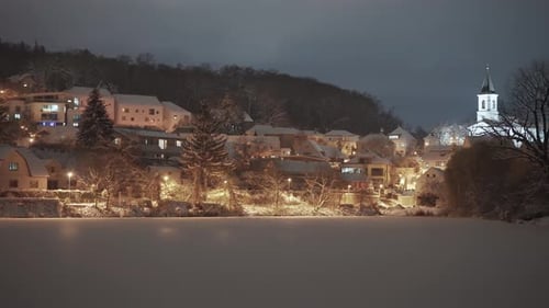 Nighttime in a quiet Prague neighborhood reveals snow-covered trees and houses, with a church standi