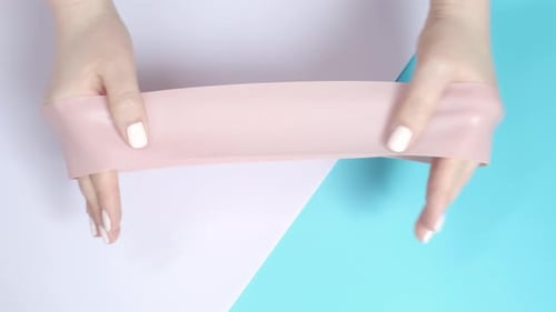 Hands stretching fitness elastic band on pink and blue background close-up.