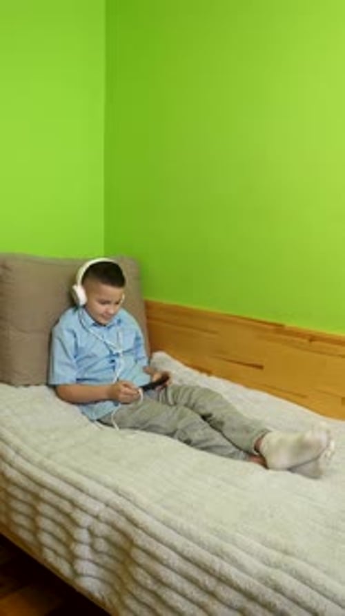 Boy With Headphones Uses Handheld Device on Bed