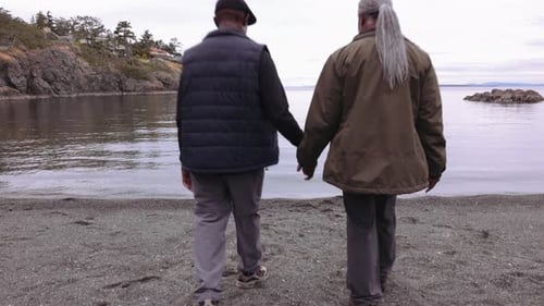 Senior Couple Holding Hands and Walking Toward Ocean on Beach 6064 Years