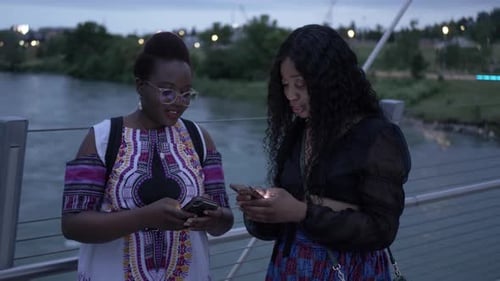 Young women friends enjoying social media on footbridge over river at night