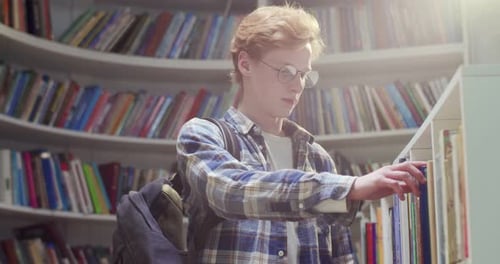 Young Redhaired Caucasian Male Student with Backpack Walking in Library Choosing Textbook to Read