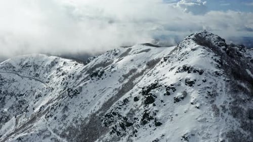 Snow-capped mountain peaks with clouds swirling around, filmed during the day, aerial view