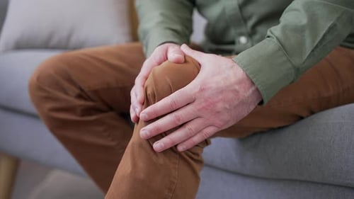Close-up of male hands massaging knee while sitting on sofa at home. Man suffering from severe knee