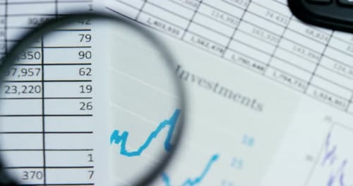 A hand with a magnifying glass passes over a financial newspaper and financial bag with a business
