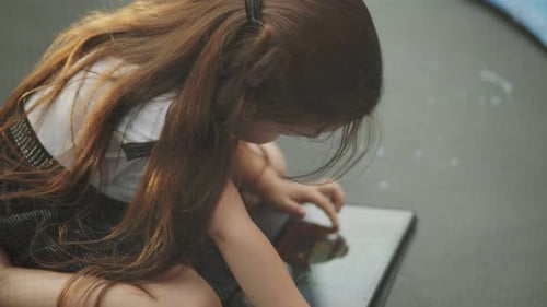 Child Sitting on a Trampoline and Looking at a Tablet