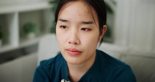 A stressed young woman sits alone on a sofa with a stressed and worried expression on her face.