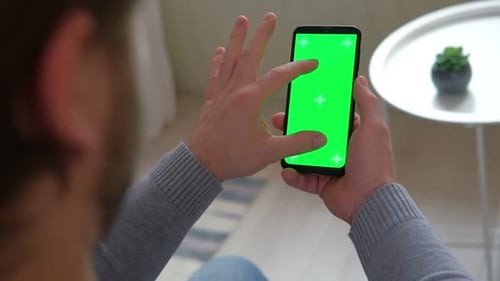 Young Man Sitting at Home Holding Smartphone Green Mockup Screen in Hand Male Person Using Chroma