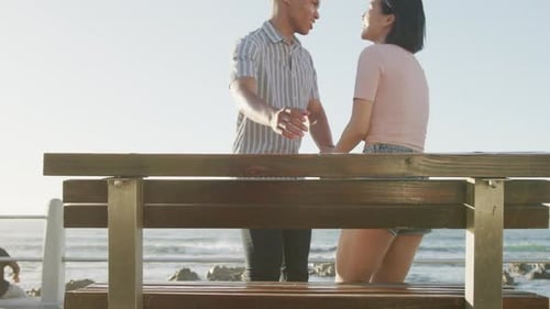Happy biracial couple sitting on bench on promenade, in slow motion