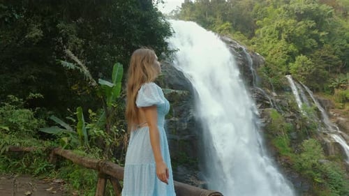 Young Woman in Blue Dress Gazes at Majestic Waterfall in Lush Green
