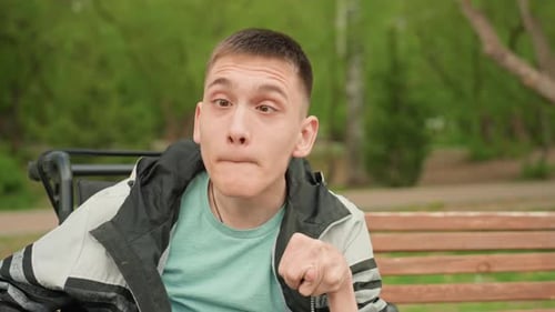 Caucasian Young Man In Wheelchair Making Exaggerated Funny Faces On Park Bench Playful Performance