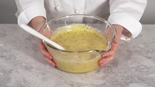 Zusammenfalten von Regenbogenstreuseln zu einem Kuchenteig in einer Glas-Rührschüssel, um Funfettti Bundt-Kuchen zu backen