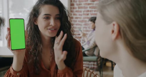 Joyful Young Lady Showing Green Screen Smartphone to Female Friend Recommending Application in Cafe