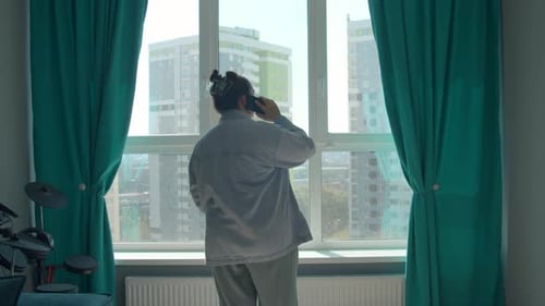 Rear view of a woman in pajamas talking on phone while standing by the window