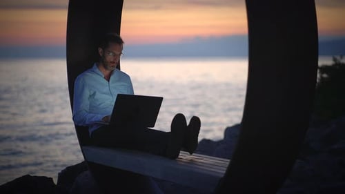 Man Working on Laptop near Ocean at Sunset
