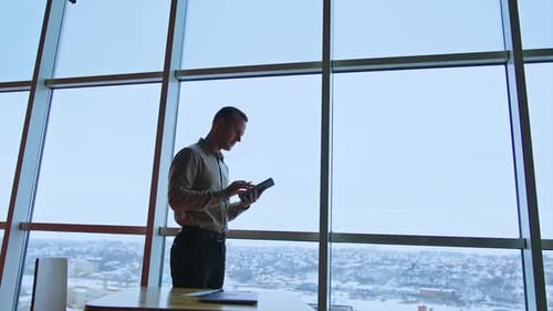 Handsome slim man stands at the window looking into his notebook. Office worker takes some notes.