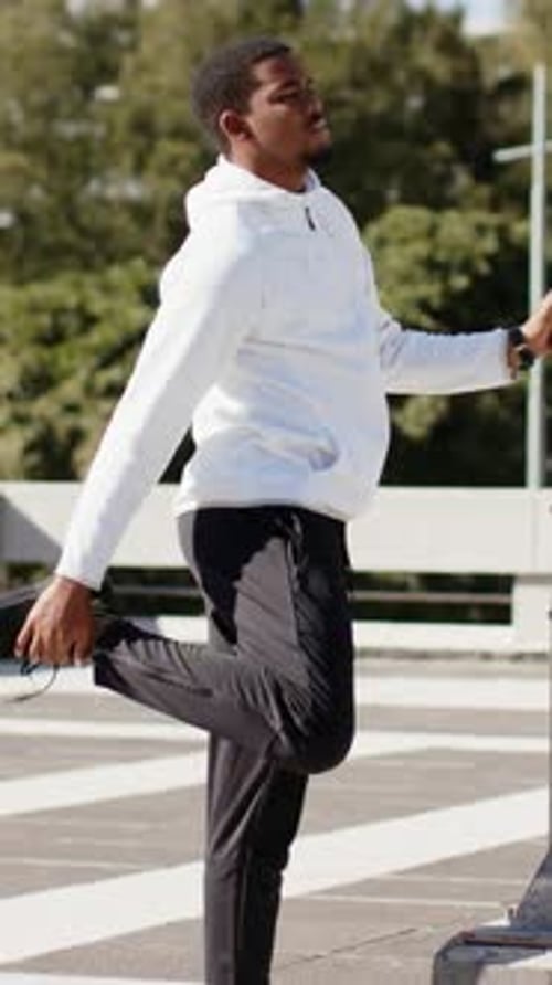 Vertical video: Walking man bracing lamppost and stretching quadriceps on rooftop, with smartwatch