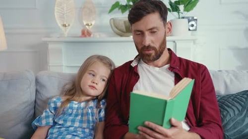 Bearded man reading book to daughter on sofa