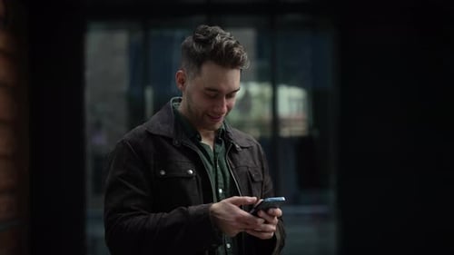 Portrait of Cheerful Young Man Standing on City Street with Smile on Face Using Smartphone to Check