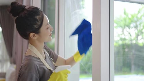 junge Frau in gelben Gummischutzhandschuhen putzt zu Hause Fenster mit Lappen und Reinigungsspray
