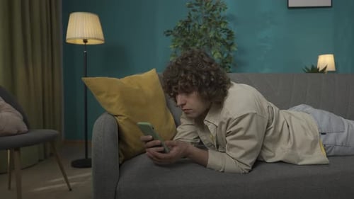 Junger Mann in Freizeitkleidung liegt mit Smartphone auf dem Sofa im Wohnzimmer und hat einen