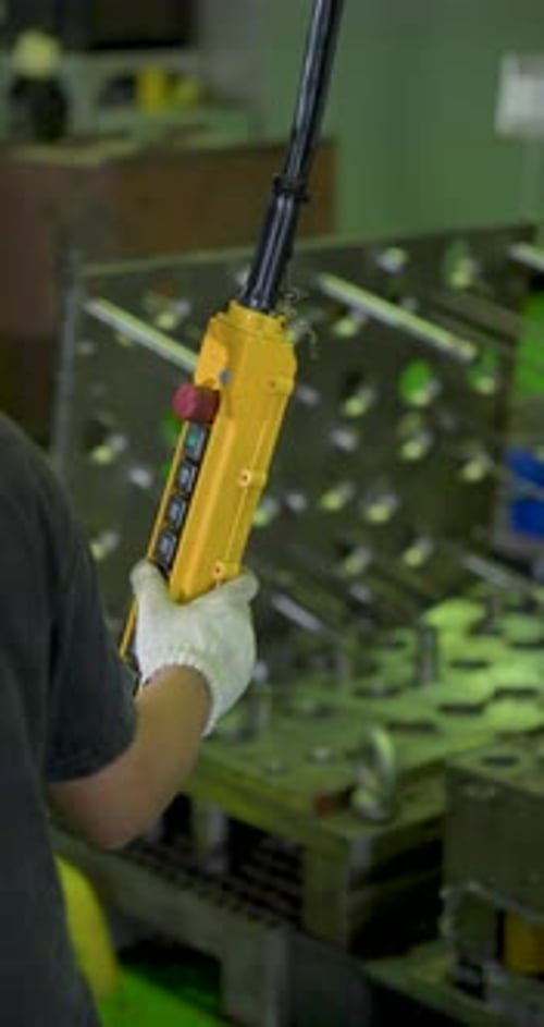 Gloved factory worker grips yellow control pendant, guiding lifting movement above tooling area