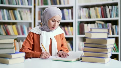 Muslim female student in hijab studies by reading books, taking notes in campus library space.