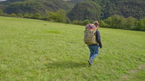 Following hiking backpacker walking down a hilly green grass meadow landscape in slow motion