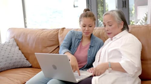 Using laptop, young asian woman teaching elderly asian woman on couch at home